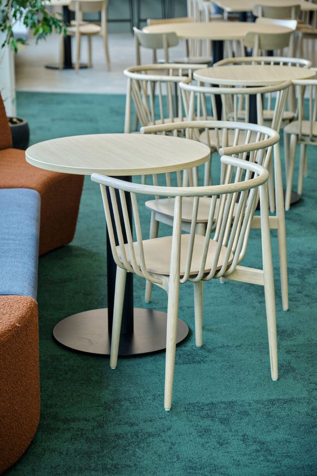 A stylish hospitality or commercial interior featuring multiple pale timber café tables with matching spindle-back chairs arranged on a teal carpet. A partial curved sofa in blue and rust upholstery appears at the left, with additional seating, tables, and indoor greenery in the background. The scene has a contemporary, warm and inviting fit-out.