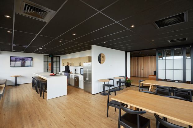 Open-plan contemporary commercial interior with a kitchenette and central white island, multiple timber dining and meeting tables, black chairs, light timber-look flooring, and a dark acoustic tiled ceiling with recessed lighting and exposed air-conditioning grilles. Glazed partitions and timber wall panelling define areas toward the rear.
