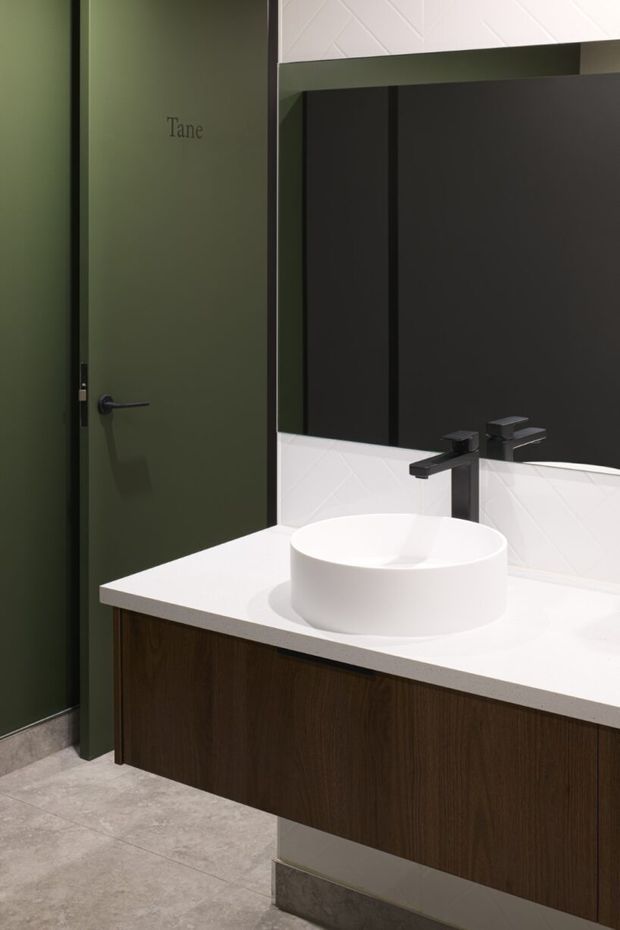 A contemporary bathroom vanity featuring a round white vessel basin, matte black wall-mounted tapware, dark timber floating cabinetry, and a white benchtop. The room has a large black-framed mirror, white geometric-patterned wall tiles, grey floor tiles, and a dark olive-green door labelled 'Tane'.