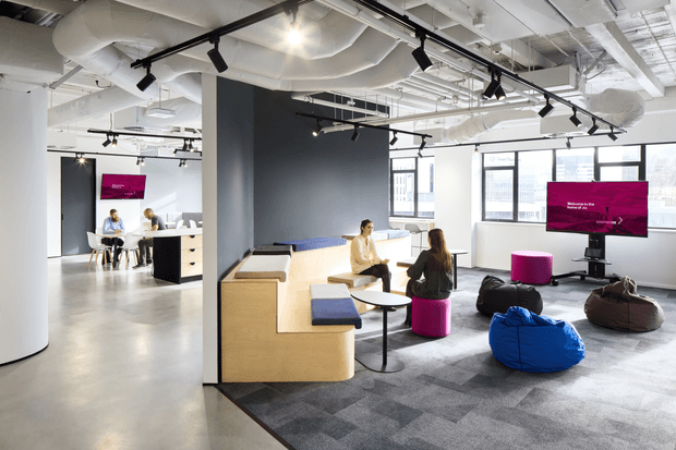 Open-plan contemporary office interior with exposed services, track lighting, large windows, collaborative booth seating, small meeting tables, beanbags, televisions, and a mix of carpeted and polished concrete flooring. The space has a bright, informal workplace atmosphere with bold magenta accents and dark feature walls.