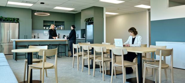 A bright contemporary commercial interior resembling a staff breakout area, office café, or shared workplace lounge. The open-plan space includes a long communal dining table with light timber chairs, a kitchen or refreshment counter with stools and appliances at the rear, and a person working on a laptop. Pale grey flooring, muted green cabinetry and walls, white acoustic ceiling tiles, and teal upholstered bench seating create a clean, calm workplace environment.