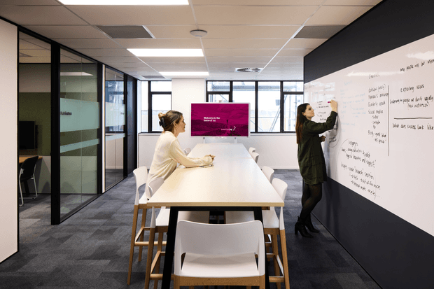 Bright contemporary commercial collaboration room with a long white meeting table, mixed white and timber chairs, dark carpet, glass-partitioned offices, and a large wall-mounted whiteboard. Two people are working at the table and writing on the whiteboard. A wall display and generous windows add to the open-plan office setting.