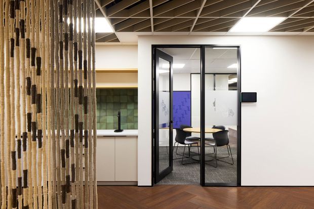 A contemporary commercial office interior with a glazed meeting room enclosed by black-framed glass doors. The space features a dark exposed-grid ceiling, white walls, herringbone timber flooring, a kitchenette or joinery zone with a green tiled splashback, and a woven rope screen in the foreground. The meeting room contains a round table and chairs, with purple acoustic wall panels visible inside.