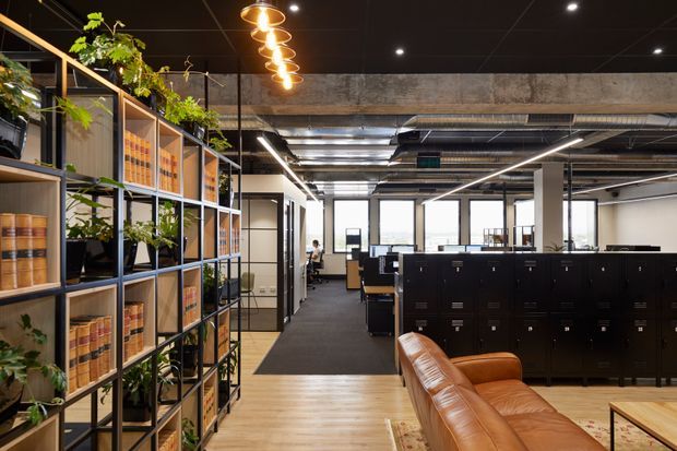 An open-plan contemporary office interior with exposed concrete and services, black acoustic ceiling, linear strip lighting, and rows of dark filing cabinets and workstations. A timber open shelving divider with integrated planter boxes separates the workspace from a lounge area featuring a tan leather sofa and timber flooring.