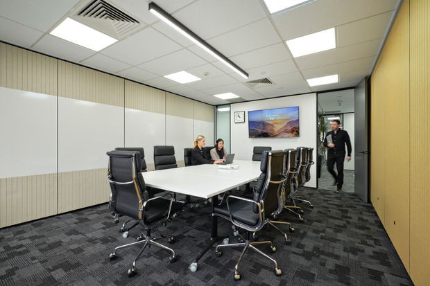 A contemporary commercial meeting room with a large white conference table surrounded by black wheeled office chairs. The room features acoustic-style timber wall panelling, white partition panels, dark patterned carpet, suspended grid ceiling tiles with recessed lighting and linear lighting, and a wall-mounted display. A glazed doorway opens to a corridor.