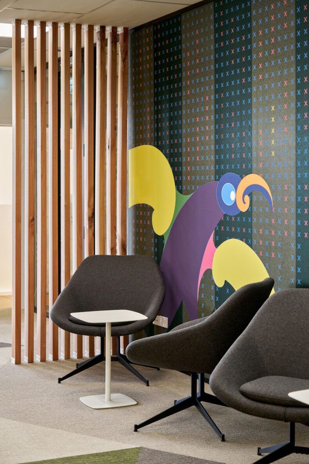 A contemporary interior lounge or waiting area with several dark upholstered lounge chairs and small white pedestal tables. A vertical timber-slat screen separates the space from an adjacent area, while the feature wall has a dark patterned surface with colourful abstract graphic shapes. Neutral carpet, exposed ceiling panels, and playful multicoloured details create a refined yet vibrant commercial-style setting.
