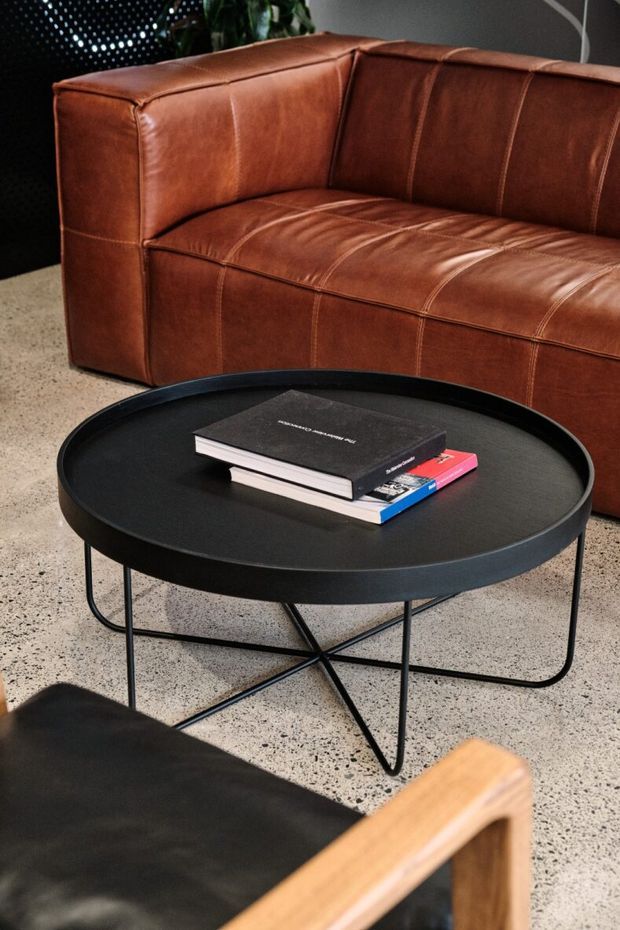 A close-up of a contemporary living room seating area featuring a cognac-brown leather sofa, a round black coffee table with a slim metal frame, stacked books, and a black leather-and-wood accent chair in the foreground. The space has a speckled polished concrete or terrazzo-like floor and a dark, refined palette.