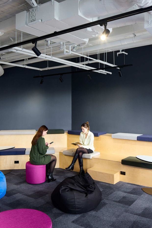 A contemporary collaborative lounge or breakout area with tiered pale timber bench seating, colourful upholstered cushions and stools, beanbag seating, and patterned carpet. The space has dark charcoal walls and an exposed services ceiling with white ducts, pipes, suspended black track lighting, and surface-mounted spotlights. Two people are seated in the informal workspace, suggesting a commercial office or creative studio environment.