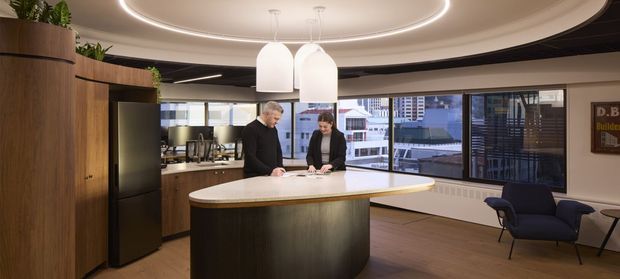 A sophisticated contemporary commercial interior featuring a curved kitchen or hospitality island with dark timber cabinetry and a light stone benchtop. Two people stand at the island beneath sculptural pendant lights and a recessed circular ceiling detail. Large windows provide an urban outlook, while lounge chairs and integrated cabinetry complete the space.