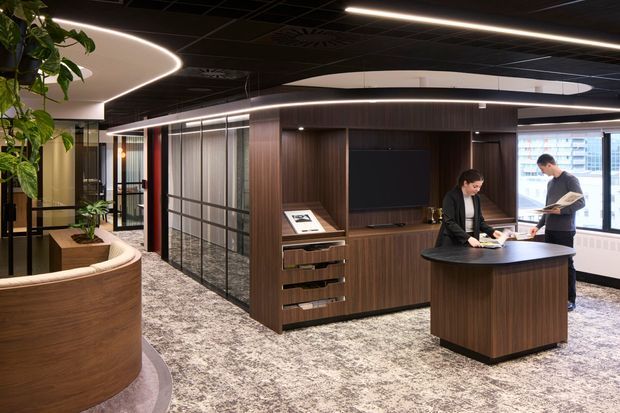 A refined contemporary commercial office interior with dark timber veneer wall panelling and built-in cabinetry, a curved reception or consultation counter, glass-partitioned work areas, integrated linear and curved LED lighting, and patterned carpet flooring. Two people are using the central workstation, while plants and city views through the windows add context.