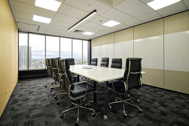A contemporary commercial conference room with a large white rectangular meeting table surrounded by black leather office chairs. The room features full-height windows with an urban view, dark patterned carpet, pale timber-toned vertical wall panelling, a white acoustic ceiling with recessed fluorescent-style lighting, and a long linear pendant light.