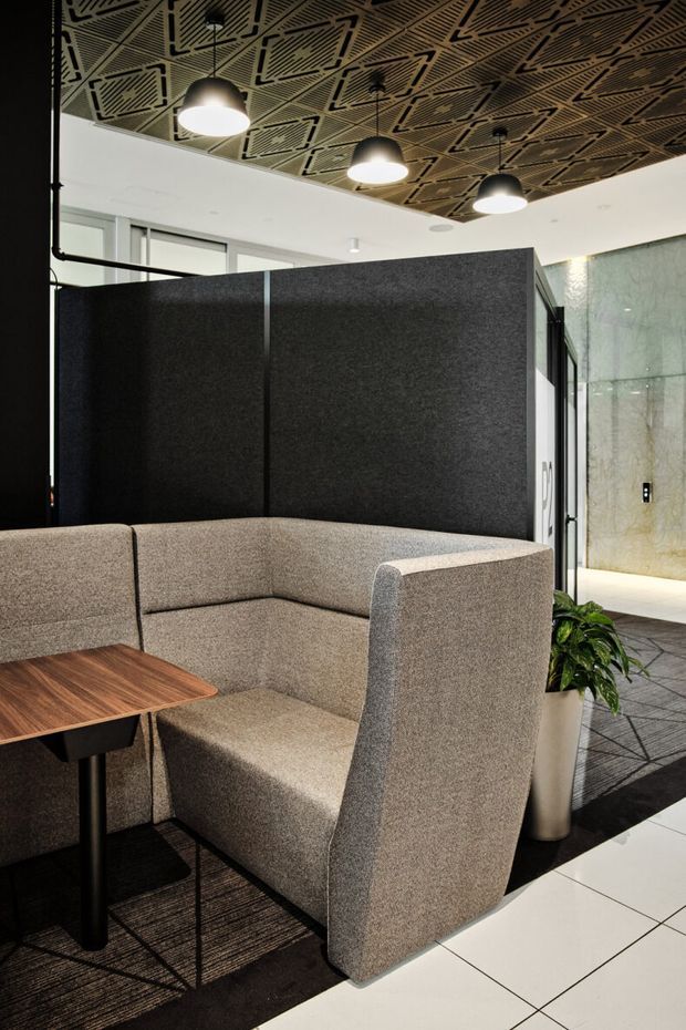 A modern commercial interior featuring a semi-enclosed upholstered booth with high grey acoustic partitions, a walnut-toned built-in table, black metal supports, and patterned acoustic ceiling panels with pendant lighting. The space also includes dark carpet, white tiled flooring, and a glazed or stone-lined area beyond the booth.