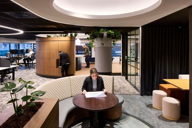 A contemporary commercial office interior with a collaborative lounge and workspaces. The foreground features a woman seated at a round dark wood table on a curved quilted banquette, with upholstered stools nearby. Behind her are open-plan desks, a rounded timber-clad meeting pod, glass-partitioned rooms, black drapery, indoor planting, patterned carpet, and large circular recessed ceiling lights. The space has a refined, professional hospitality-inspired atmosphere.
