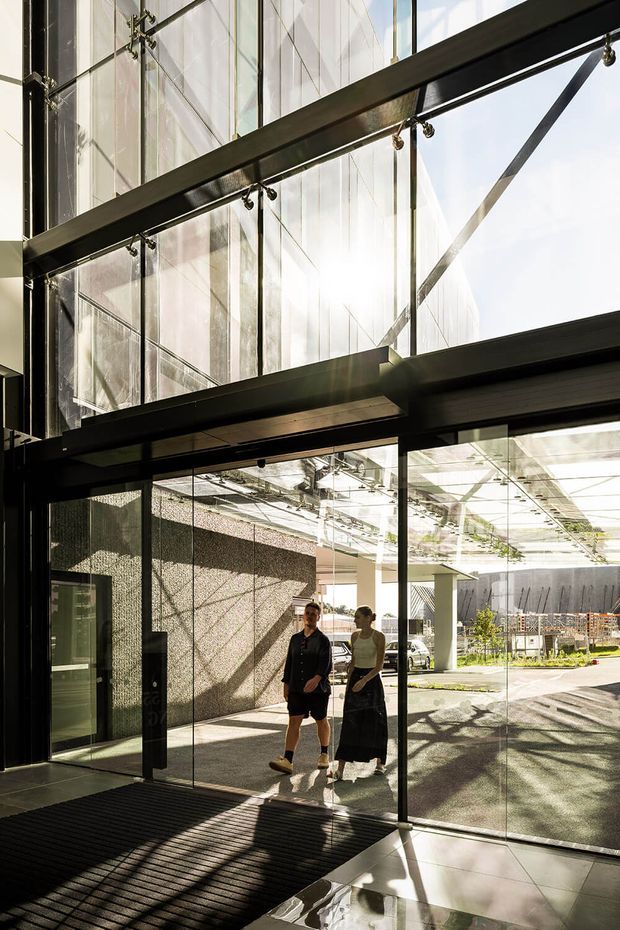 A dramatic double-height glazed entry or lobby with extensive floor-to-ceiling glass, exposed dark steel structural beams, and large framed glass doors. Strong sunlight casts geometric shadows across the dark entrance mat and polished light floor. Two people stand near the transparent entrance, with an outdoor courtyard and modern building elements visible beyond.