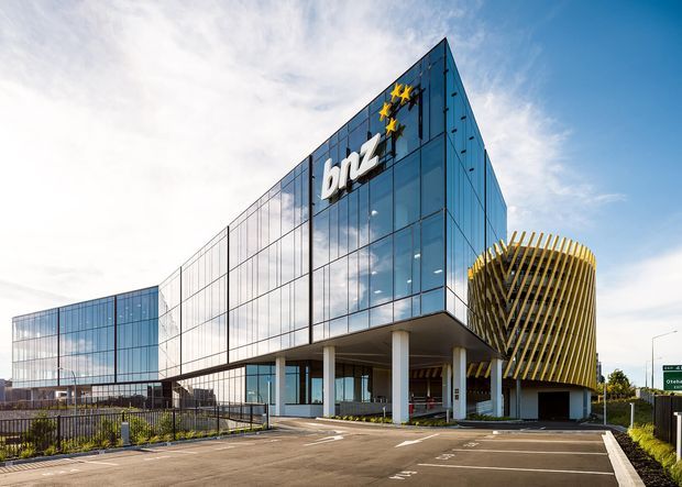 Exterior architectural photograph of a contemporary commercial building, featuring a sharply angled glazed facade with prominent BNZ branding and a distinctive cylindrical volume wrapped in vertical gold-toned fins. The building is elevated on white columns above a paved parking and drop-off area, with extensive curtain-wall glazing and a clear sky.