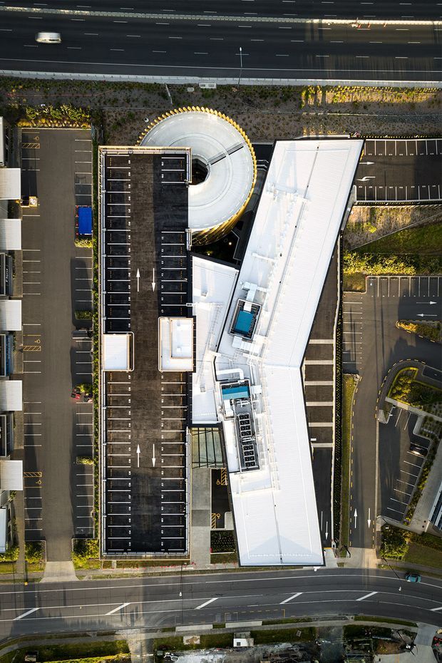 Aerial, near-vertical view of a large contemporary commercial building complex with bright white angular roof forms, rooftop mechanical equipment, a circular rooftop structure, extensive surface parking, landscaped planted strips, and surrounding multi-lane roads. The image is primarily an architectural site and building-form study viewed from above.