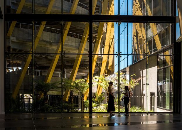 Interior view of a large contemporary glazed atrium or commercial lobby, looking through floor-to-ceiling curtain walls into a planted courtyard. Bold yellow structural braces, exposed steel framing, concrete surfaces, reflective glazing, and strong sunlight define the space. Two people provide scale, while the polished dark floor and layered architectural reflections add a dramatic, high-contrast atmosphere.