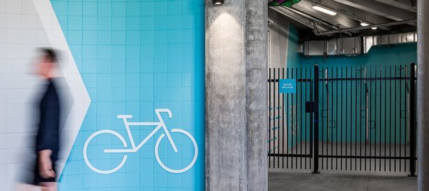 An urban commercial interior or building entry area with a vivid turquoise tiled wall featuring a large white bicycle graphic, a raw concrete structural column, exposed ceiling services, and a black metal security gate. A blurred pedestrian appears at the left edge. The space has an industrial, utilitarian character with strong graphic wayfinding or branding.