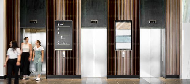 Interior view of a contemporary elevator lobby with multiple elevator doors set into dark stone or metal surrounds and vertical timber-look slatted wall panels. Lift indicators, floor directory signage, and call buttons are visible, with people standing near the elevators. The space has a polished, commercial architectural fit-out with a neutral grey, black, and warm wood palette.
