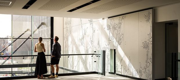 A bright contemporary interior circulation space or landing with full-height glazing, a glass balustrade, and a perforated white architectural screen wall. Two people stand by the windows, with an urban structure visible beyond. The ceiling has recessed linear lighting and exposed ventilation grilles.