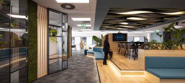 Contemporary commercial office interior with an open-plan hospitality and breakout area. The space features glazed meeting rooms, vertical timber slats, a living green wall, patterned carpet, timber flooring, teal upholstered banquette seating, a kitchen or café counter with bar stools, abundant indoor planting, and a sculptural layered ceiling feature. The image has a polished, professional workplace atmosphere with strong zoning between circulation and social spaces.