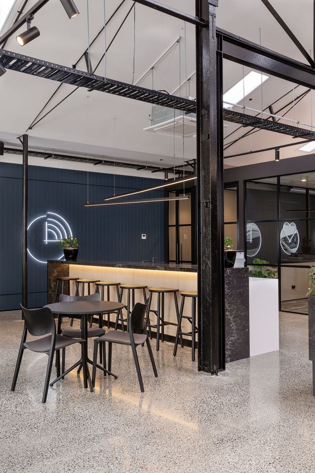 A contemporary commercial café or breakout area within an industrial-style office interior. The space features a long illuminated bar with stools, small dining tables and chairs, dark vertical wall panelling, black steel structural columns, exposed cable trays and services, suspended linear lighting, internal glazed meeting-room partitions, and a polished speckled concrete or terrazzo floor. A neon-style circular sign and indoor plants add visual accents.