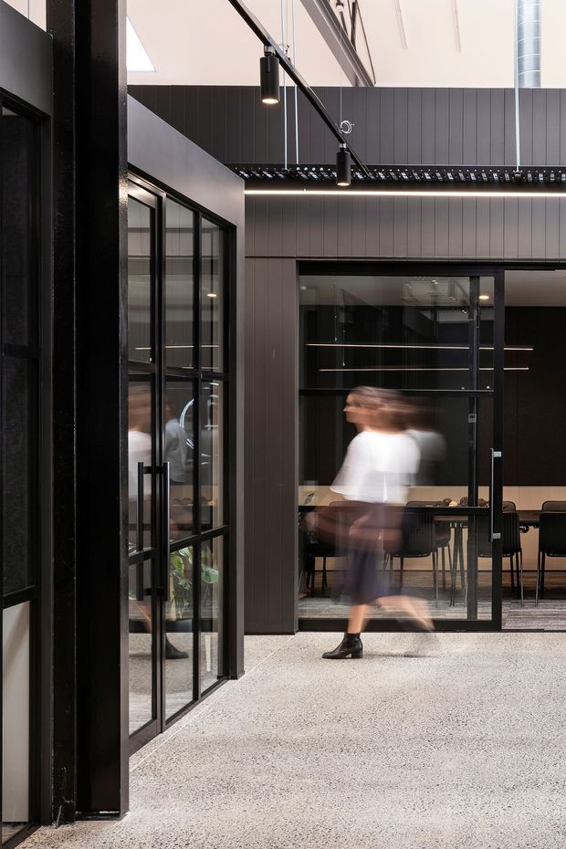 A contemporary commercial interior with a dramatic double-height atrium or circulation space. Black steel-framed glazed partitions and doors define meeting rooms, with dark vertical wall panelling, exposed structural elements, linear pendant lighting, and a polished speckled floor. A dining or meeting area is visible through the glazing, while motion-blurred people emphasize the active workplace atmosphere.