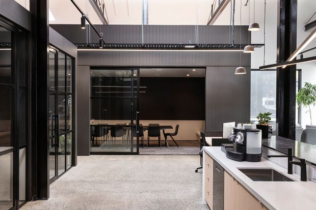 Contemporary open-plan office interior with a compact kitchenette, black-framed glass partitions, a meeting room, high ceilings, exposed dark structural elements, and pendant lighting. The space combines light cabinetry and benchtops with dark wall cladding and a carpeted floor.