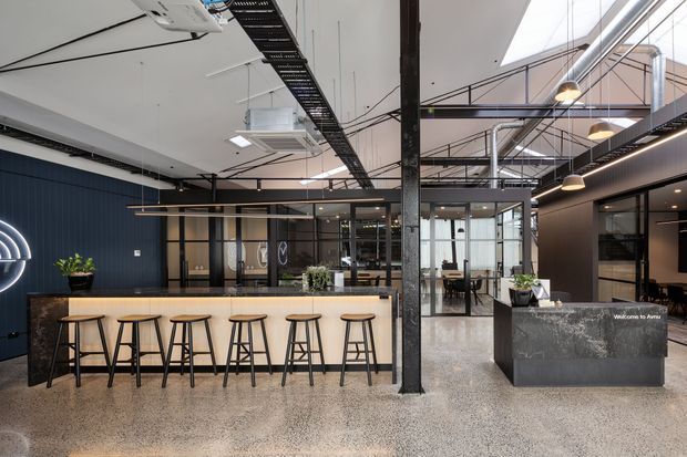 A spacious contemporary commercial interior featuring a polished concrete floor, a long timber-fronted bar or kitchenette with black stools, and a dark stone reception counter. The space has exposed steel columns, visible services and cable trays, black-framed glazed meeting rooms, pendant lighting, and large skylights, creating an industrial office or showroom atmosphere.