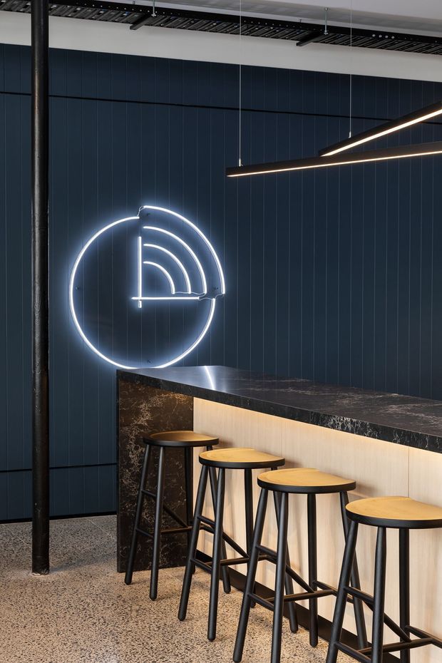 A contemporary commercial bar or reception counter with a dark vertically panelled wall, illuminated circular logo, black marble-look countertop, pale timber-toned front, and a row of black stools with round timber seats. Linear pendant lighting, exposed dark ceiling services, a black structural column, and a polished terrazzo-style floor create an industrial, refined atmosphere.