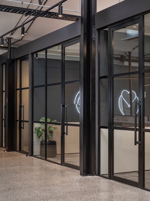 A contemporary commercial interior corridor lined with black-framed glazed partitions and doors. The space features exposed black ceiling services and suspended linear lighting, dark acoustic wall panels, polished concrete or terrazzo-style flooring, and a potted plant. Glass reveals private office or meeting areas with a neon-style illuminated graphic.
