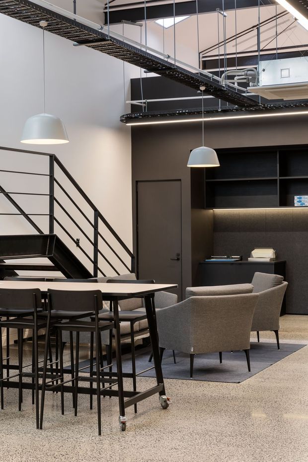 A sophisticated open-plan commercial interior with a dining or meeting area in the foreground and a lounge/reception area beyond. The space features polished terrazzo-style flooring, charcoal built-in cabinetry, a dark metal staircase and exposed overhead cable trays and services. Grey upholstered chairs, a long table, pendant lights, linear LED lighting, and a high ceiling create a refined industrial office or hospitality atmosphere.