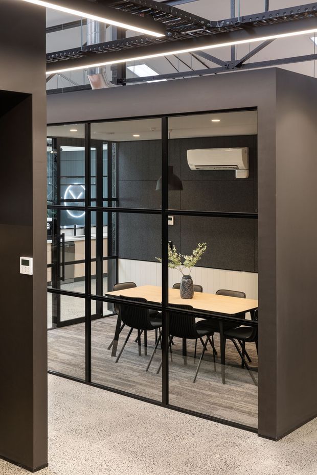 Modern commercial meeting room enclosed by black-framed glass partitions, with a light timber meeting table, black chairs, dark acoustic wall panels, and a wall-mounted split-system air conditioner. The space opens to an industrial-style office area with exposed services, cable trays, ductwork, and structural elements overhead. Polished speckled flooring and linear LED lighting complete the fit-out.