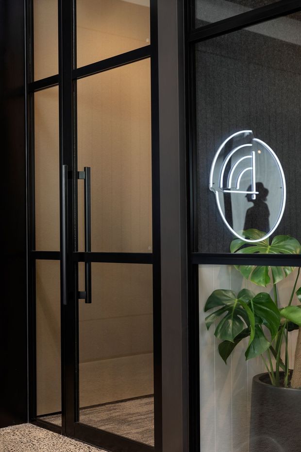 Close-up view of a dark-framed glazed entry door and adjacent internal glazing, featuring a long vertical pull handle, warm translucent panels, a circular illuminated wall sign or reflection, and a large potted monstera plant. The scene has a refined, modern commercial or hospitality character with terrazzo flooring and subdued lighting.