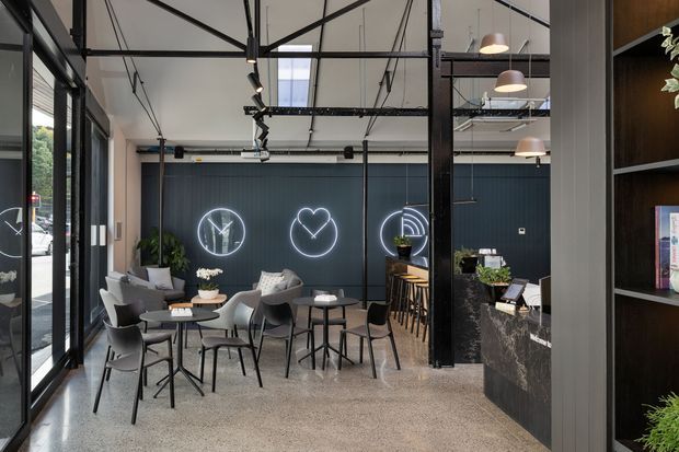 A contemporary commercial café or hospitality interior with an open-plan seating area, polished concrete floor, dark charcoal feature wall with illuminated graphic signage, exposed black structural framing and services, pendant lighting, indoor plants, and a stone-clad service counter with timber shelving. Full-height glazing opens the space visually to the street.