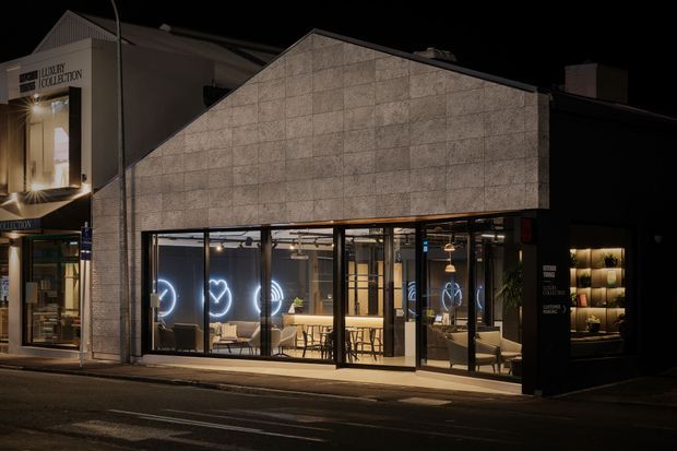 Night-time street view of a contemporary commercial shopfront or showroom. The building features a large pitched/gabled facade clad in pale textured panels, extensive full-height dark-framed glazing, and a brightly illuminated interior with pendant lights, tables, chairs, shelving, and display cabinetry. Adjacent retail signage is visible, but the main subject is the renovated commercial facade and glazed interior.