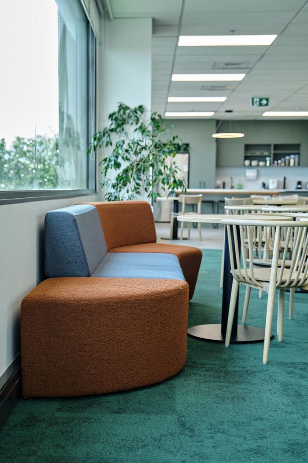A bright contemporary office lounge and dining area with floor-to-ceiling windows, a colourful upholstered modular lounge chair, round café tables, pale timber-framed chairs, teal carpet, a kitchenette or breakout counter, indoor plants, and suspended and recessed ceiling lighting. The space has an open-plan commercial workplace character.