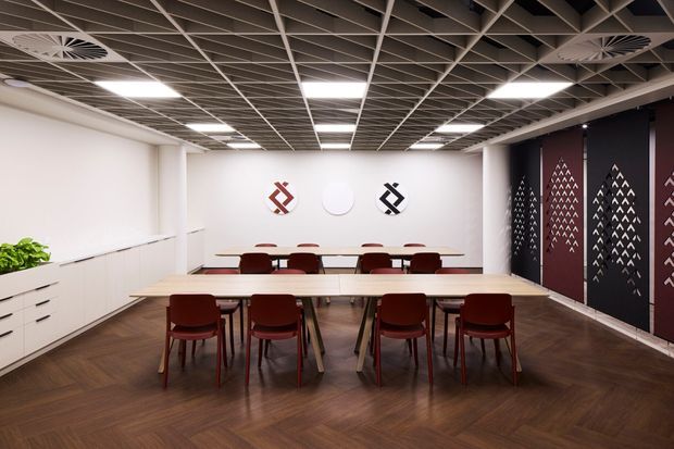 A contemporary commercial meeting or collaborative workspace with long white tables, red upholstered chairs, white built-in storage, and a dark patterned feature wall. The room has a dark wood-look floor, suspended open-grid ceiling with recessed rectangular lights and circular air diffusers, and a clean, corporate interior aesthetic.