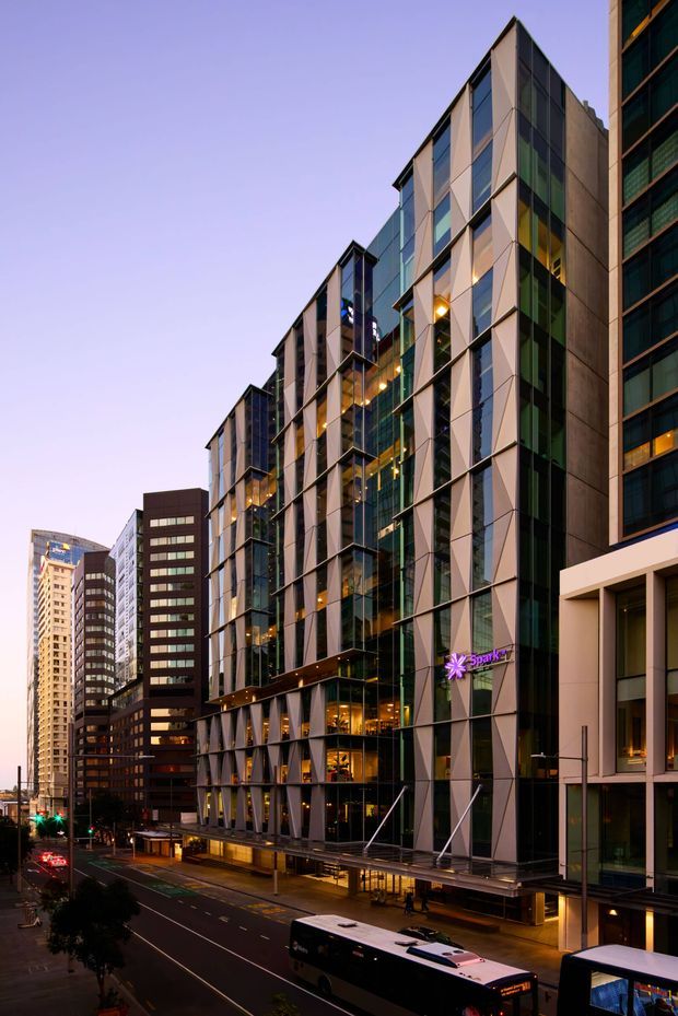 Twilight urban streetscape featuring a tall contemporary commercial building with a highly glazed facade, angular projecting fins, dark metal framing, and illuminated ground-floor retail or lobby spaces. Dense high-rise buildings line the street, with buses and traffic lanes in the foreground. The scene has a polished metropolitan character with warm interior lighting contrasting against the cool dusk sky.