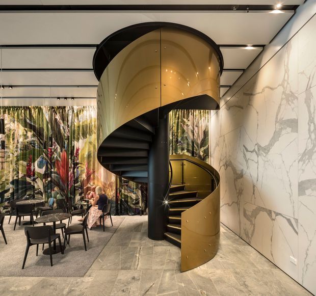 A dramatic contemporary interior featuring a sculptural black-and-brass spiral staircase beneath a curved metallic canopy. The space includes large marble-look wall panels, pale stone-look flooring, dark ceiling beams, patterned floor-to-ceiling curtains, and a small seating area with black chairs and tables. Warm integrated lighting highlights the staircase and creates a refined, luxurious atmosphere.