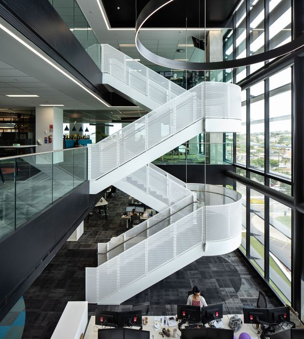 A large, contemporary multi-level commercial interior dominated by a sculptural white zigzag staircase with perforated metal balustrades and curved landings. The stair rises through a tall glazed atrium with extensive floor-to-ceiling windows, black structural framing, suspended circular lighting, glass balustrades, and open-plan workstations below. Dark carpet, black ceilings, and white stair elements create a high-contrast, professional office environment.
