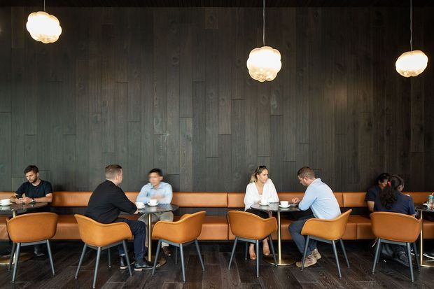 A contemporary commercial dining or café interior with a long built-in cognac leather banquette against a dramatic dark timber-panelled feature wall. Several small round tables are paired with orange upholstered dining chairs, while sculptural pendant lights hang overhead. The space has dark timber flooring, a warm low-key atmosphere, and is occupied by diners.