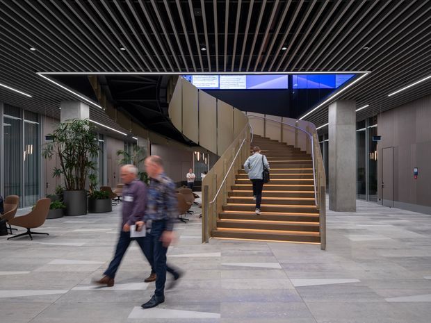 A contemporary commercial interior lobby or atrium with a broad sculptural staircase as the central feature. The space has a dark linear slatted ceiling with recessed lighting, pale grey large-format floor tiles, concrete structural columns, glazed and panelled walls, integrated seating, indoor planting, and illuminated stair nosings. People are moving through the lobby, which appears to be a modern workplace or public building circulation space.