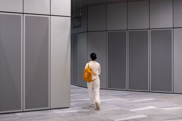 A modern interior passage or lobby with large modular grey wall panels, perforated metal screening, and a pale stone or concrete-look tiled floor. A person walks toward a suspended directional sign, suggesting a commercial or institutional building such as a gallery, convention centre, or office complex. The space is minimalist, monochromatic, and softly lit.