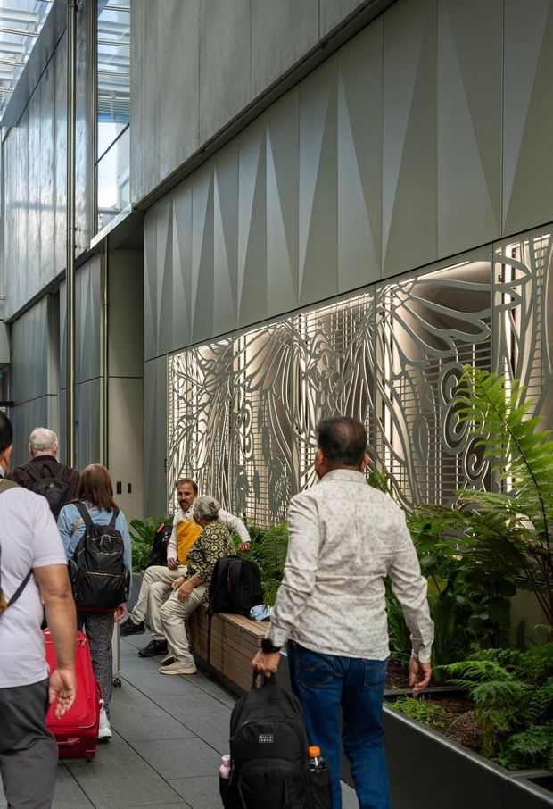A tall, contemporary indoor public passage or commercial atrium with a glazed roof, exposed concrete walls, grey metal wall panels featuring faceted geometric relief, and an intricate perforated metal decorative screen. Built-in planter beds with lush greenery line the walkway, which is occupied by pedestrians and luggage. The space has strong vertical proportions, abundant natural light, and an urban architectural character.