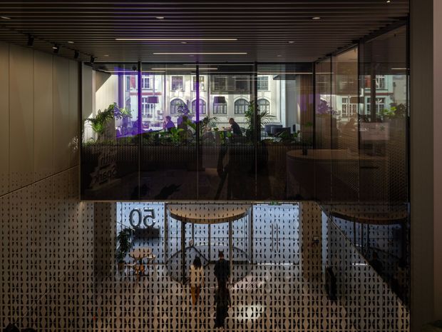 Dark, atmospheric view into a contemporary commercial interior through extensive glazing. A perforated decorative screen forms the foreground balustrade or partition, with a planted workspace or lounge beyond. The ceiling has dark linear timber or slatted panels with recessed and track lighting, while reflections and the view outside reveal an urban setting and neighbouring buildings.