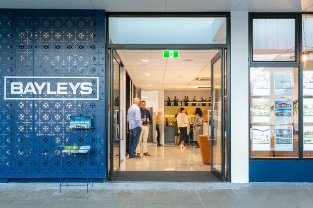 Contemporary commercial interior entrance with a prominent blue decorative perforated feature wall displaying the BAYLEYS logo. The glazed doorway opens into a bright reception or office area with people, shelving, polished flooring, and warm timber accents. Large framed windows and property marketing displays flank the entrance.