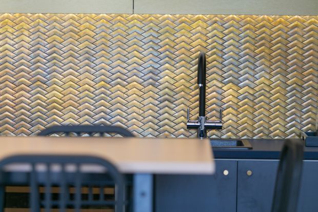 Close-up view of a contemporary kitchen featuring dark blue lower cabinetry, a black gooseneck tap, and a striking gold and grey chevron-patterned tiled splashback. The foreground includes a blurred dining table and chairs.