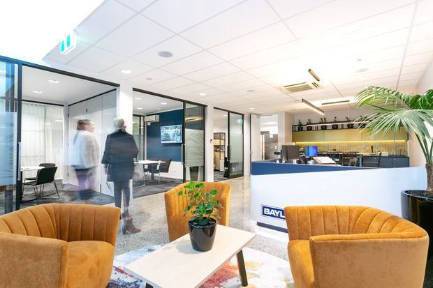 Bright contemporary commercial reception and lounge area with mustard upholstered armchairs, a light coffee table, glass-partitioned meeting rooms, a reception desk, and a compact kitchenette or refreshment counter. The space has a suspended acoustic tile ceiling, polished stone or terrazzo-look flooring, black-framed glazing, and indoor planting.