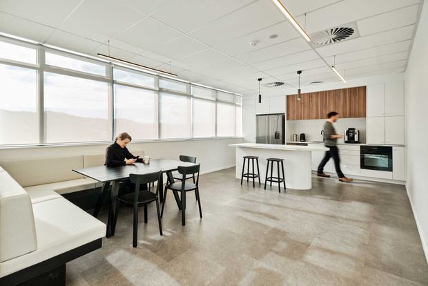 Bright open-plan office breakout area combining a small dining/work table, white built-in bench seating, and a compact kitchenette with island breakfast bar. The space has expansive windows, a white suspended grid ceiling with linear lighting, pale flooring, and minimalist white cabinetry accented by a vertical timber feature panel.